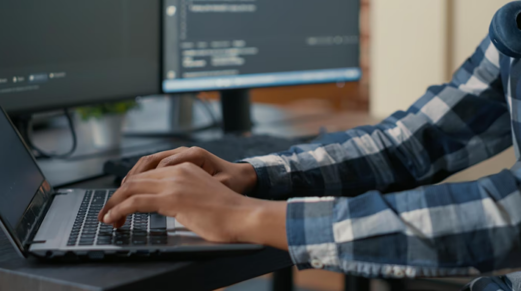 man in blue shirt typing on laptop keyboard and two computer screens behind